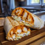 A cross-section of a spicy buffalo chicken wrap, showing chunks of orange-coated chicken and creamy coleslaw inside a tortilla.