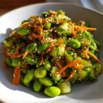Close-up of a vibrant edamame salad featuring bright green edamame, chopped cucumber, shredded carrots, and toasted sesame seeds.