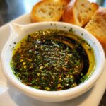 Close-up of a white bowl containing a flavorful bread dip made of olive oil, balsamic vinegar, and mixed herbs.