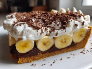 Close-up of a slice of banoffee pie showing layers of crust, chocolate, bananas, whipped cream, and chocolate shavings.