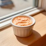 A small white ramekin filled with creamy, orange-hued homemade burger sauce sitting on a wooden surface near a window.