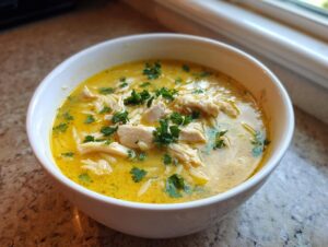 Close-up of a white bowl filled with rich, yellow chicken orzo soup, topped with shredded chicken and fresh parsley.
