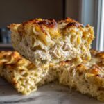 Close-up of a thick slice of creamy chicken pasta bake being lifted, showing shredded chicken and a browned, cheesy top.