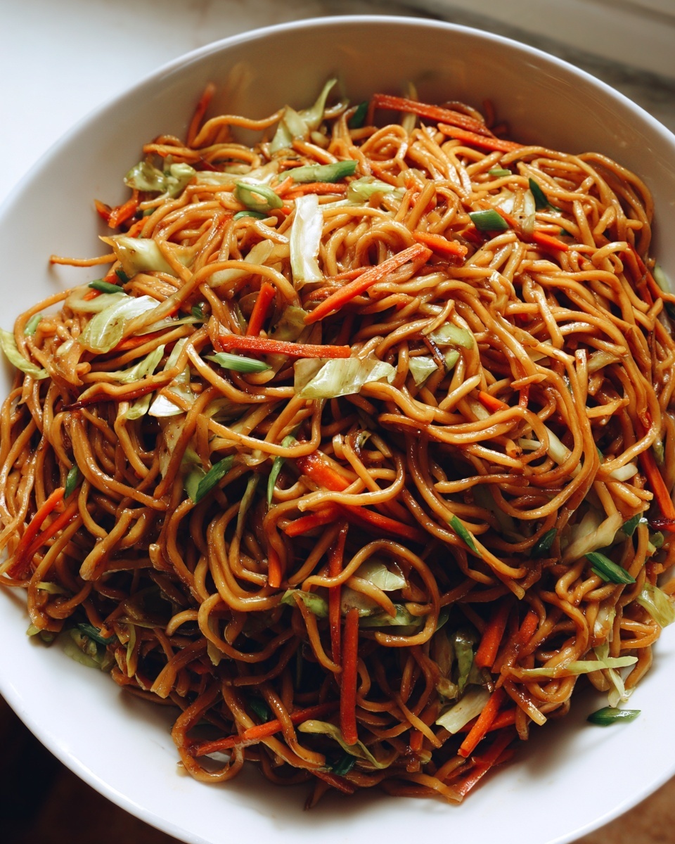 A close-up overhead shot of a large white bowl filled with savory, saucy fried noodles mixed with shredded carrots and cabbage.