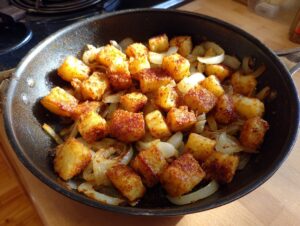 Close-up of golden brown, crispy fried potatoes mixed with caramelized onions cooking in a black skillet.