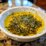 Close-up of a small white bowl filled with golden olive oil bread dip topped with minced garlic and herbs.