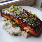 Close-up of a perfectly cooked piece of teriyaki salmon with a dark, glossy glaze, topped with sesame seeds and green onions, resting on white rice.