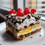 A perfect slice of banana split cake featuring layers of crust, fruit, cream, and topped with whipped cream, chocolate drizzle, and maraschino cherries.