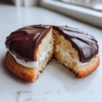 A close-up of a black and white cookie cut in half, showing the cake, cream filling, and chocolate glaze.