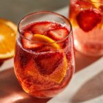 Close-up of a glass filled with bubbly red champagne punch, garnished with sliced strawberries and an orange wedge.