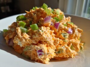A close-up mound of vibrant orange buffalo chicken salad topped with chopped green onions and red onion pieces.