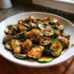 A close-up of a white bowl filled with glistening chicken zucchini stir fry, garnished with sesame seeds and scallions.