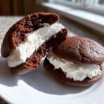 Two rich chocolate whoopie pies, one cut in half to show the thick, fluffy white cream filling.