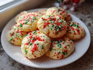A white plate stacked with soft, crinkled christmas sprinkle sugar cookies topped with red, green, and white holiday sprinkles.