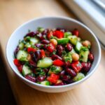Close-up of a white bowl filled with a colorful dense bean salad featuring kidney beans, cucumber, and red pepper.