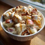 A white bowl filled with creamy apple salad featuring chunks of red-skinned apple, marshmallows, pecans, and drizzled with caramel sauce.