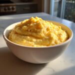 A white bowl filled with creamy, bright yellow cheese grits, sitting on a sunlit marble countertop.