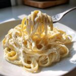 A fork lifting fettuccine noodles coated in thick cottage cheese alfredo sauce, sprinkled with pepper.