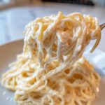 Close-up of creamy, cheesy protein pasta being lifted high on a fork from a plate.