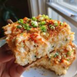 A hand holding up a thick slice of creamy sushi bake, topped with green onions and togarashi spice.