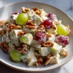 A close-up serving of creamy waldorf salad featuring chunks of apple, red and green grapes, and toasted walnuts.