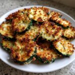 A white plate piled high with crispy, golden-brown baked parmesan zucchini slices.