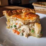 Close-up of a generous slice of chicken pot pie recipe showing creamy filling, shredded chicken, peas, and carrots under a flaky crust.