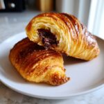 A flaky, golden brown chocolate croissant cut in half, revealing melted chocolate filling inside.