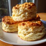 A close-up of three tall, fluffy buttermilk biscuits stacked on a white plate, showing golden tops.