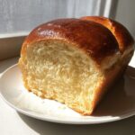 A freshly baked, glossy loaf of condensed milk bread sliced to show its soft, fluffy interior on a white plate.