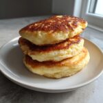 A stack of three thick, golden-brown greek yogurt pancakes served on a light ceramic plate.