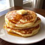 Close-up of a tall stack of fluffy pancakes topped with melting butter and drizzled with syrup.