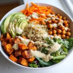 Close-up of a colorful buddha bowl featuring quinoa, roasted chickpeas, avocado, and sweet potatoes drizzled with tahini dressing.