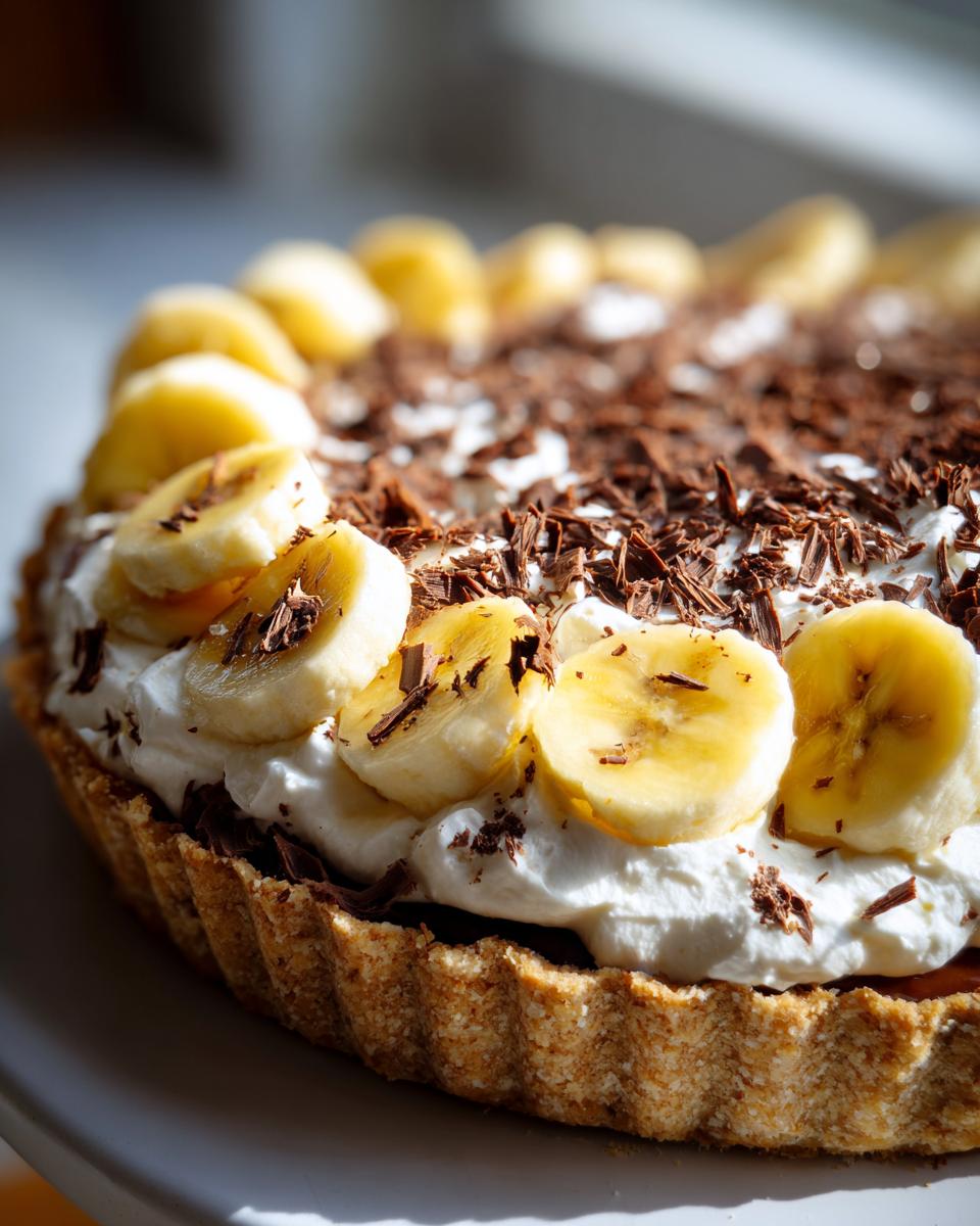 A close-up view of a homemade banoffee pie topped with whipped cream, sliced bananas, and chocolate shavings.