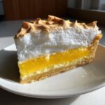 A tall slice of lemon meringue pie showing layers of crust, bright yellow curd, and fluffy, browned meringue.