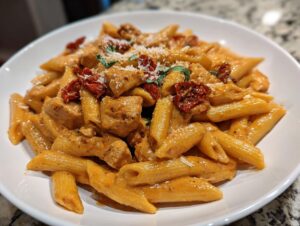 Close-up of a bowl of marry me chicken pasta featuring penne, chicken pieces, and sun-dried tomatoes in a creamy sauce.