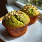 Close-up of two vibrant green pistachio muffins topped with chopped pistachios and coarse sugar.