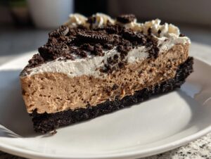 A rich slice of no-bake dirt cake featuring a dark cookie crust, light brown mousse filling, white whipped topping, and crushed cookies.