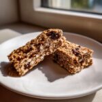 Two textured, homemade no-bake protein bar halves resting on a white plate near a window.