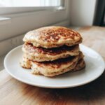 A close-up stack of four fluffy oatmeal pancakes served on a white plate near a window.