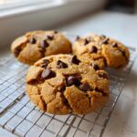 Three freshly baked peanut butter chocolate chip cookies cooling on a wire rack, topped with chocolate chips and sea salt.