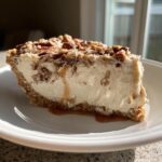 A thick slice of pecan cream pie with a graham cracker crust, topped with pecans and drizzled with caramel.