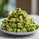 A close-up of bright green pistachio puppy chow mixed with chopped pistachios and whole almonds piled on a white dish.