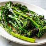 Close-up of vibrant green, roasted broccolini spears glistening with oil and seasoning on a white serving dish.