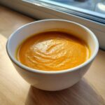Close-up of vibrant orange roasted butternut squash soup in a white bowl, sitting on a wooden surface.
