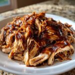 Close-up of tender, shredded crockpot teriyaki chicken smothered in glossy teriyaki sauce on a white plate.