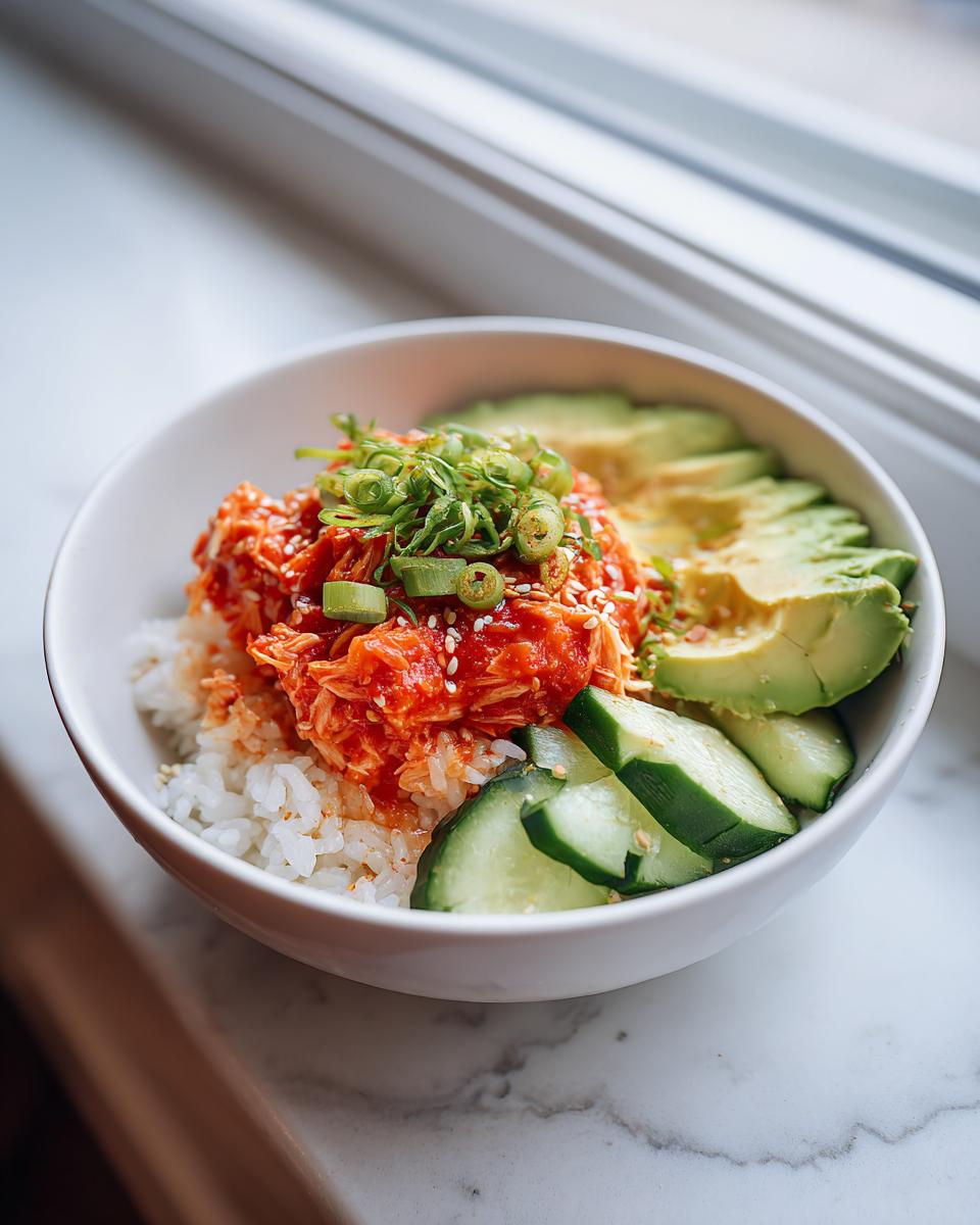 A close-up of a spicy crab sushi bowl featuring rice topped with bright orange spicy crab mix, sliced avocado, and cucumbers.