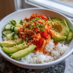 A close-up of a spicy crab sushi bowl featuring rice, shredded spicy crab mix, bright orange tobiko, sliced avocado, and cucumbers.
