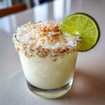 Close-up of a white Christmas margarita topped with crushed ice and toasted coconut flakes, garnished with a lime wheel.