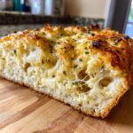 Close-up of a thick slice of cheese bread showing melted, stringy cheese and herbs on a wooden cutting board.